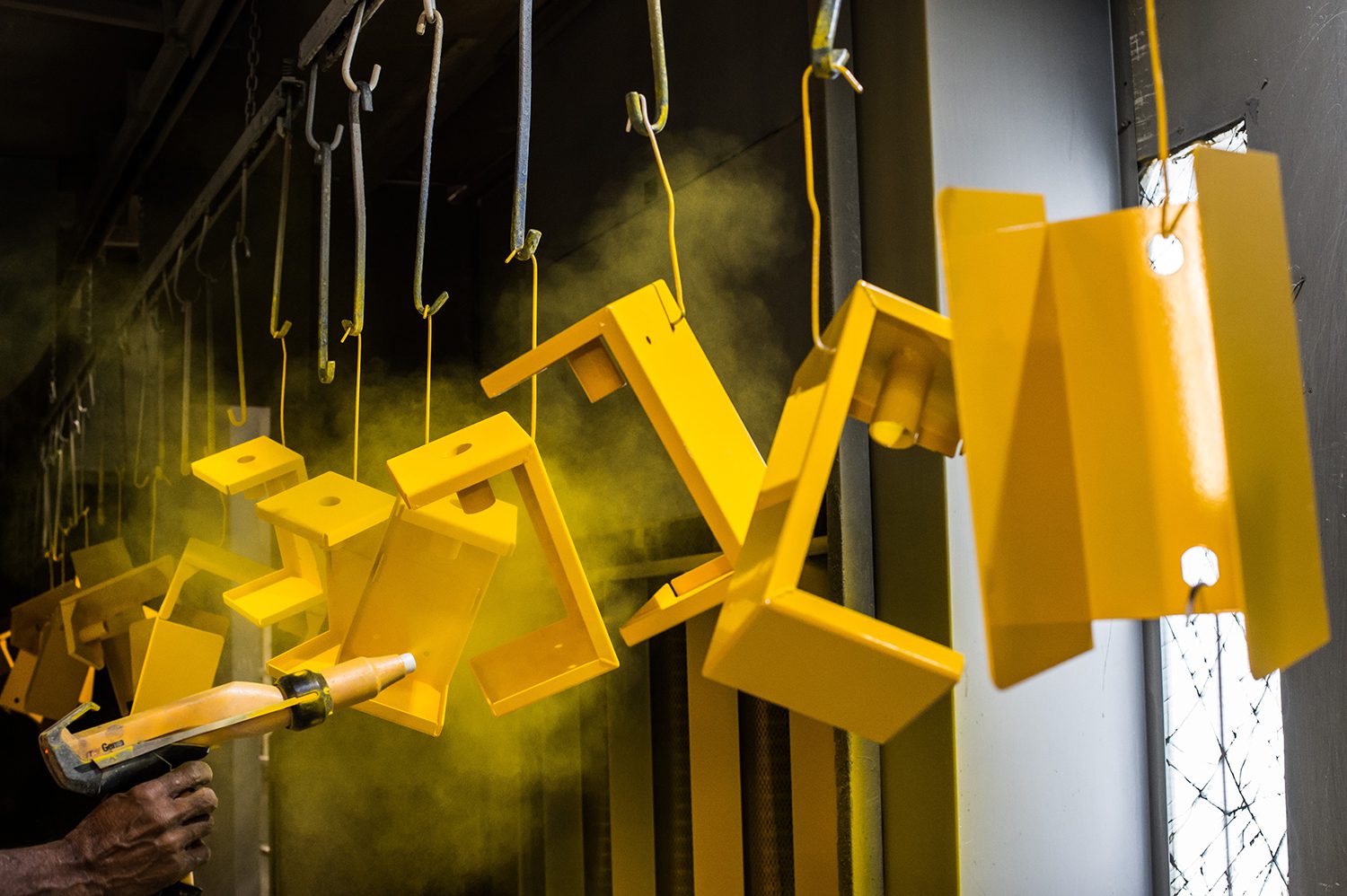 A line of metal parts hung from hooks as they are sprayed with yellow powder coating.