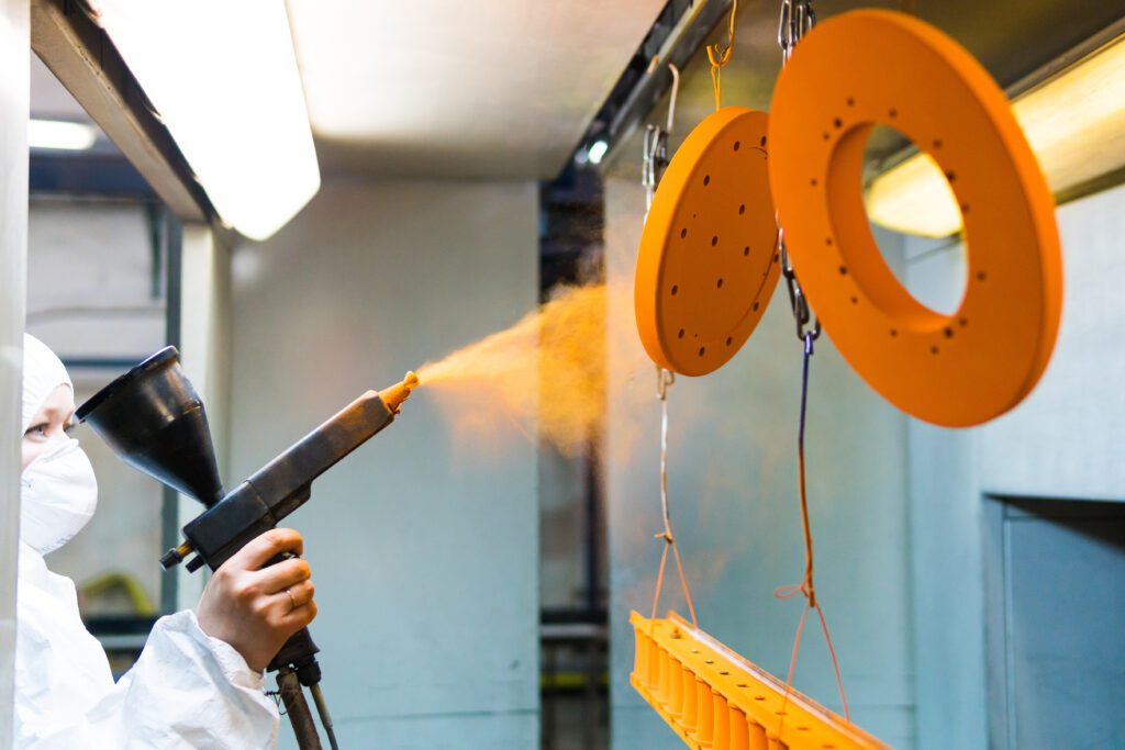 Technician in protective gear using a spray gun to powder coat orange metal parts in an industrial paint booth.