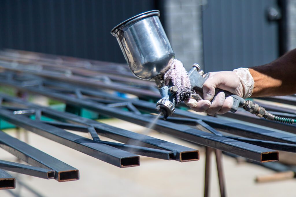 Worker using a spray gun to paint metal frames outdoors, wearing a glove for protection during the coating process.